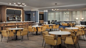 Un moderno vestíbulo de hotel con mesas y sillas redondas, una zona de bar y un área de estar con sofás.