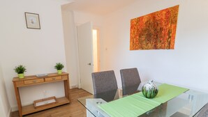 Un coin repas comprenant une table en verre, deux chaises, une petite table d’appoint en bois agrémentée de plantes et un tableau accroché au mur.