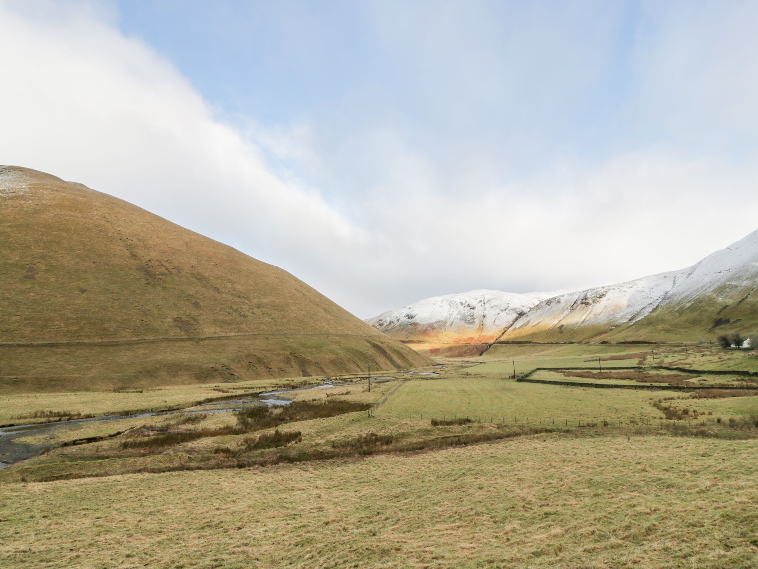 A rural landscape with a river, fields, and snow-capped hills.