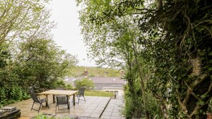 Een terras met een tafel en stoelen, omgeven door weelderige begroeiing en een uitzicht op een huis in de verte.