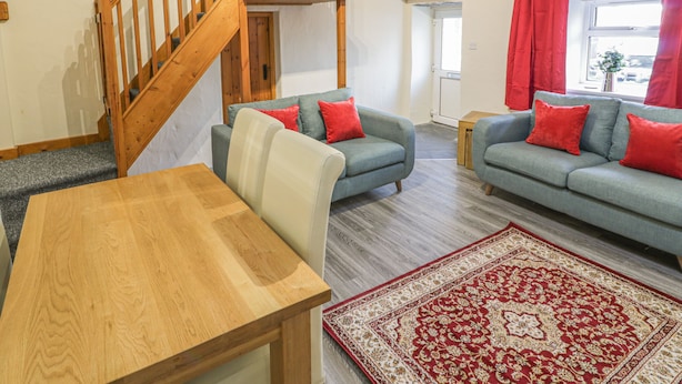Een gezellige woonkamer met een houten trap, twee banken met rode kussens, een houten eettafel en een kamerbreed tapijt met een patroon.