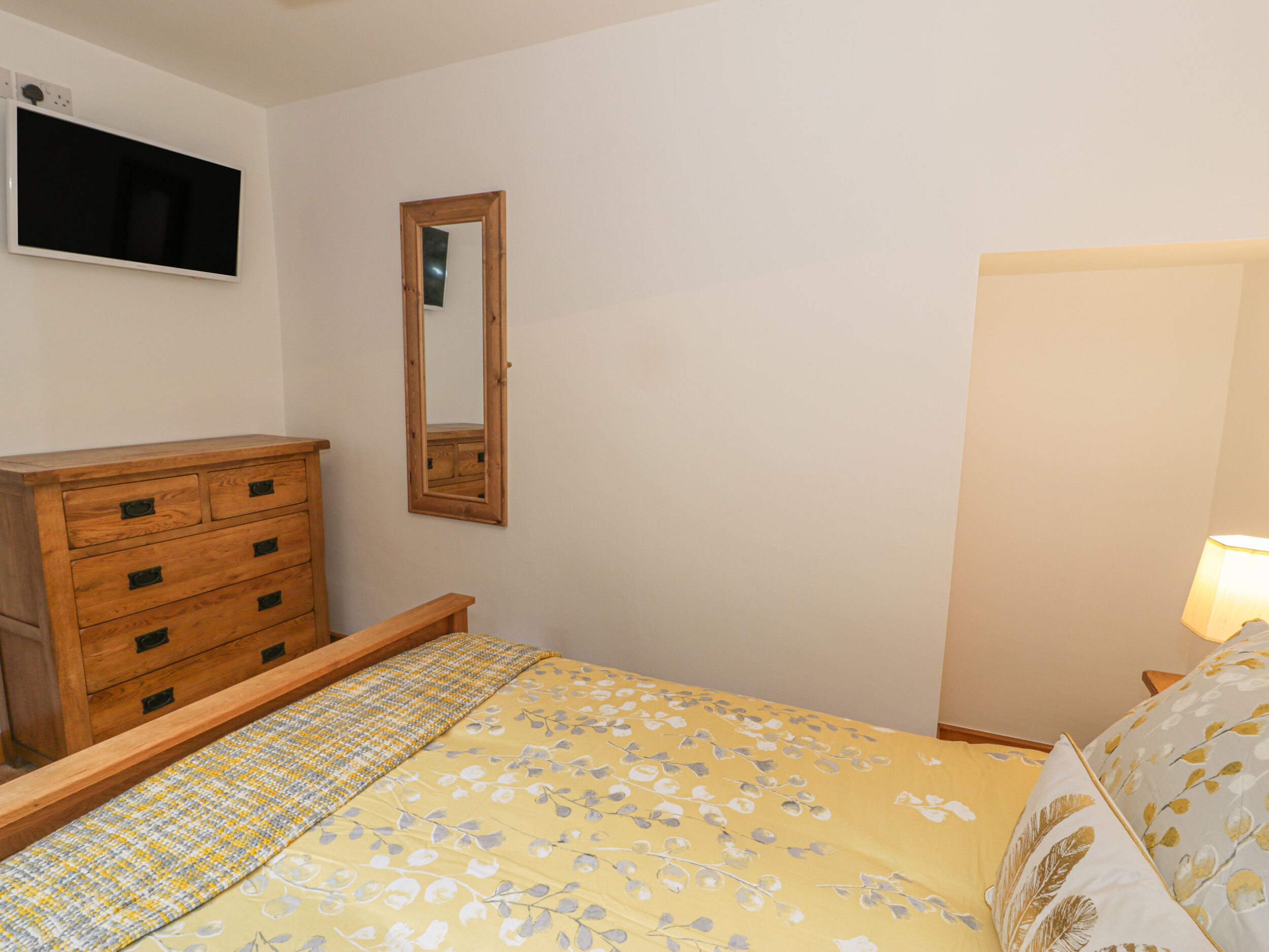 A bedroom with a wooden dresser, a bed with a floral bedspread, a mirror, and a bedside lamp.