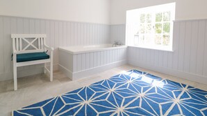 A bathroom with a white bathtub, a white bench with a teal cushion, and a large window with a view of greenery. - Carriage House (Presteigne)