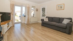 A living room with a grey sofa, a television on a wooden stand, and a door leading to a balcony. - Seagull Cottage (Holyhead)