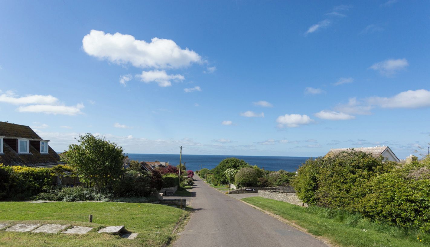 A narrow road flanked by houses with a clear view of the sea in the distance.
