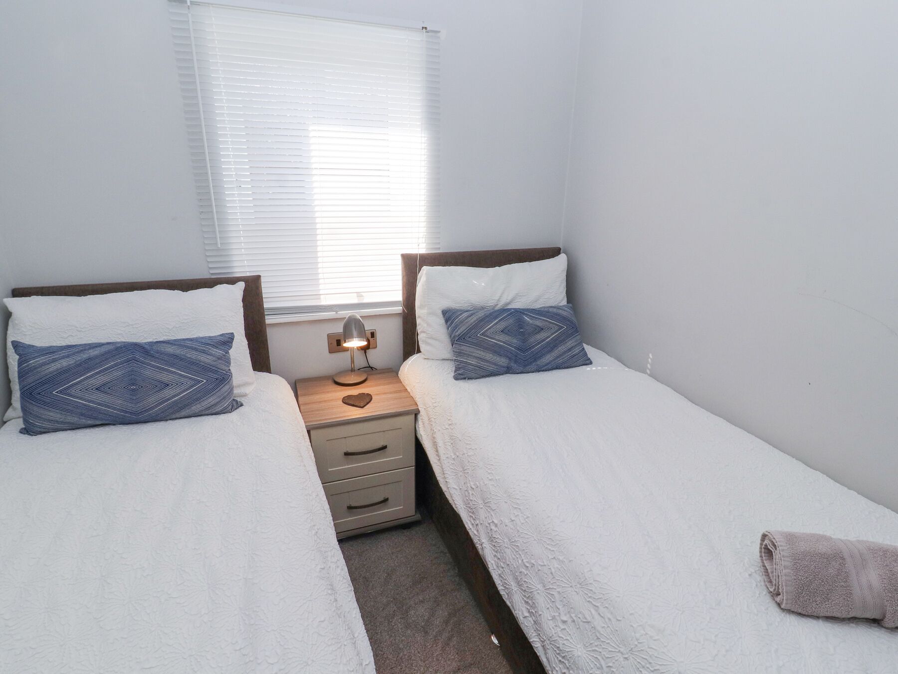A twin room with two beds, a nightstand with a lamp, and a window with blinds.