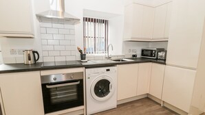 Una cocina moderna con armarios blancos, una campana extractora de acero inoxidable, horno empotrado, lavadora, microondas y una ventana con persianas.