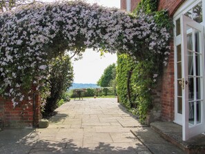 Un chemin menant à un bâtiment doté d’une porte blanche, entouré d’un mur de briques et recouvert de plantes grimpantes en fleurs.