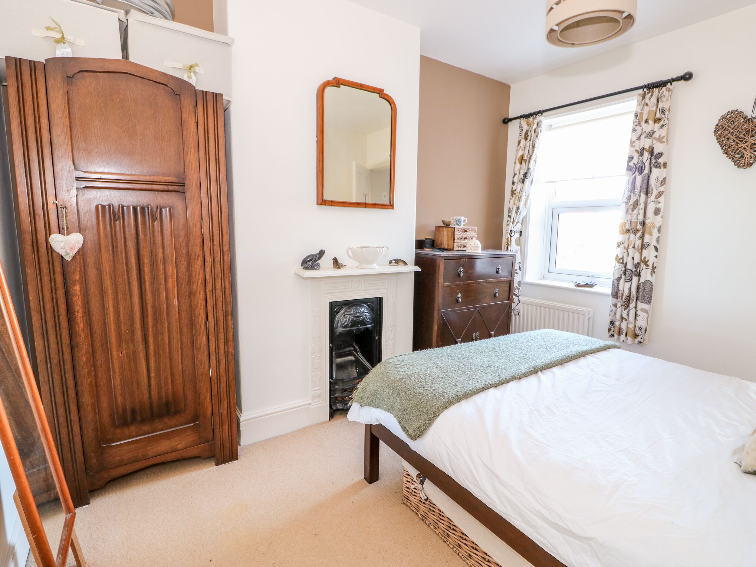 A bedroom with a wooden wardrobe, a mirror, a dresser, a bed with a green throw, and a window with patterned curtains.
