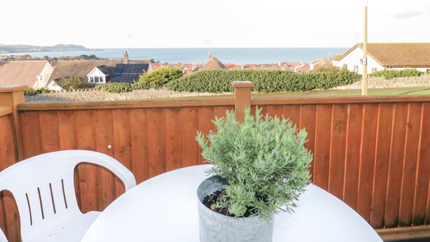 A wooden deck with a potted plant, white plastic chairs, and a view of the sea and buildings in the distance. - Bay View House (Colwyn Bay)