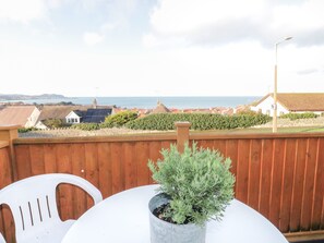 A wooden deck with a potted plant, white plastic chairs, and a view of the sea and buildings in the distance. - Bay View House (Colwyn Bay)
