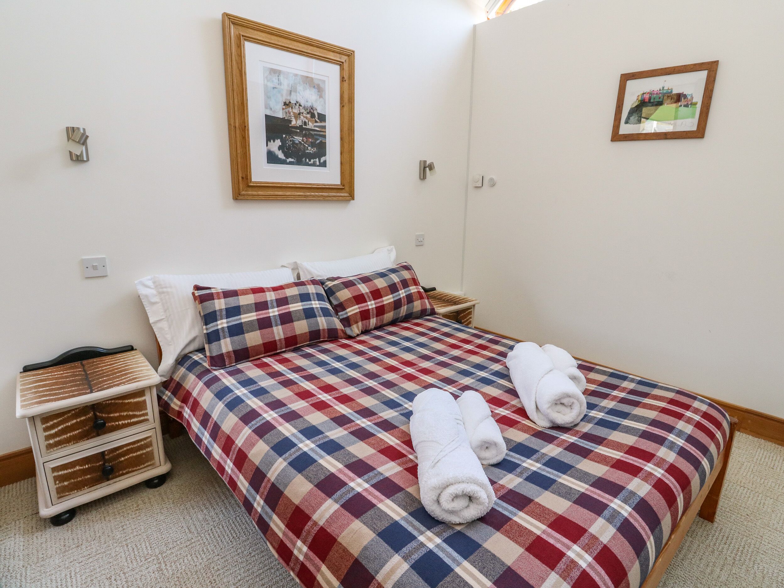 A neatly made bed with a plaid bedspread, two pillows, and rolled towels on the bed. There is a small bedside table with a lamp and a framed picture on the wall above the bed.