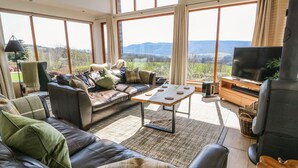 Un salón espacioso con sofá de cuero, una mesa de centro y televisor. Amplias ventanas ofrecen vistas al exterior.