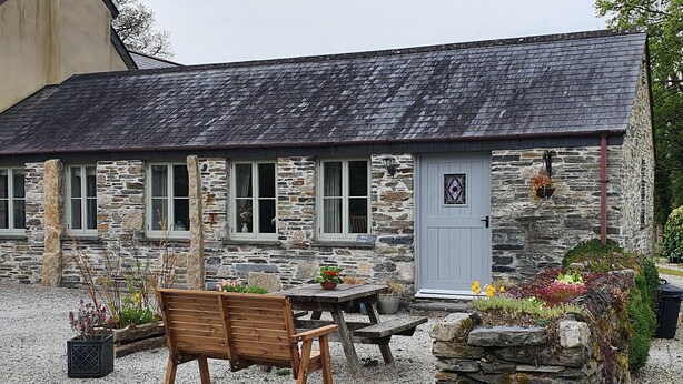 A stone cottage with a tiled roof, a wooden bench, and a table in the gravel courtyard. - Mimi's Cottage (Liskeard)