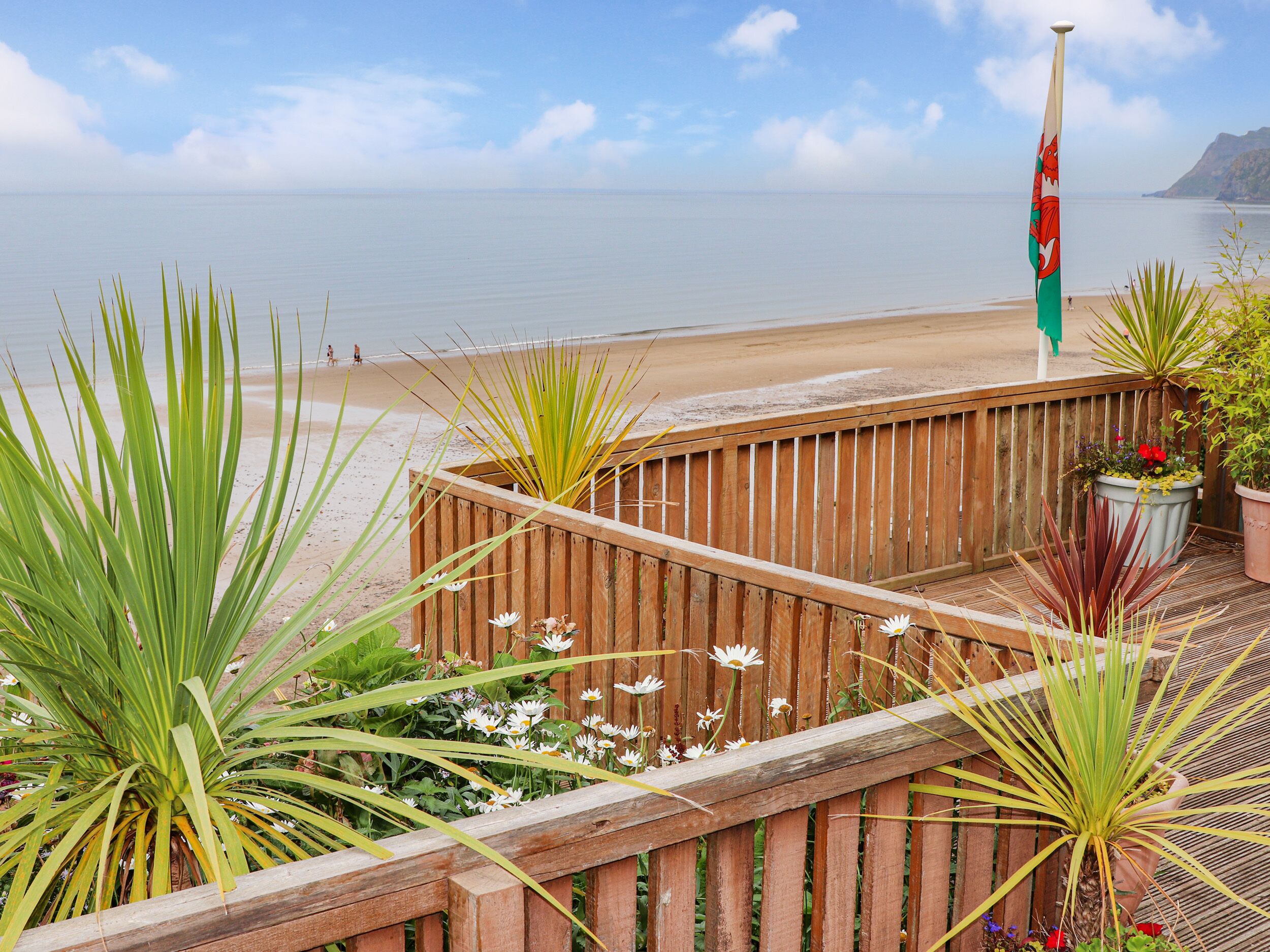 A wooden deck with potted plants and a flag, overlooking a beach and calm sea.
