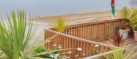 A wooden deck with potted plants and a flag, overlooking a beach and calm sea.