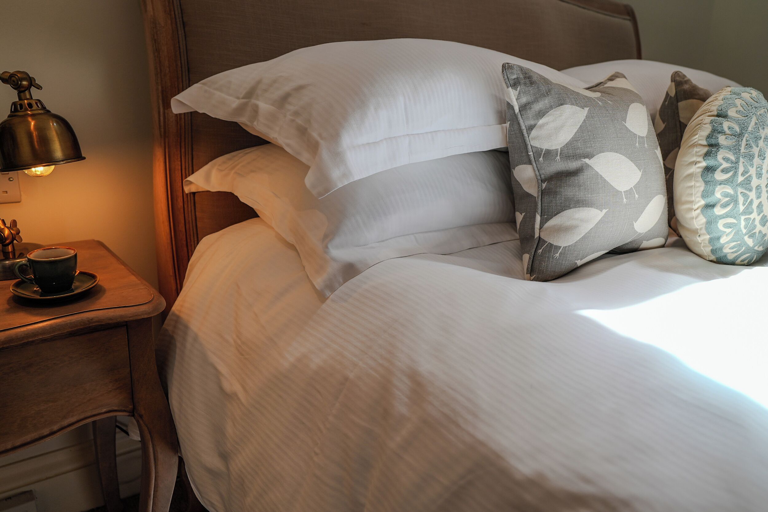 Una cama bien hecha con sábanas blancas y varios almohadones, una mesita de noche con una lámpara y una taza sobre un platillo.