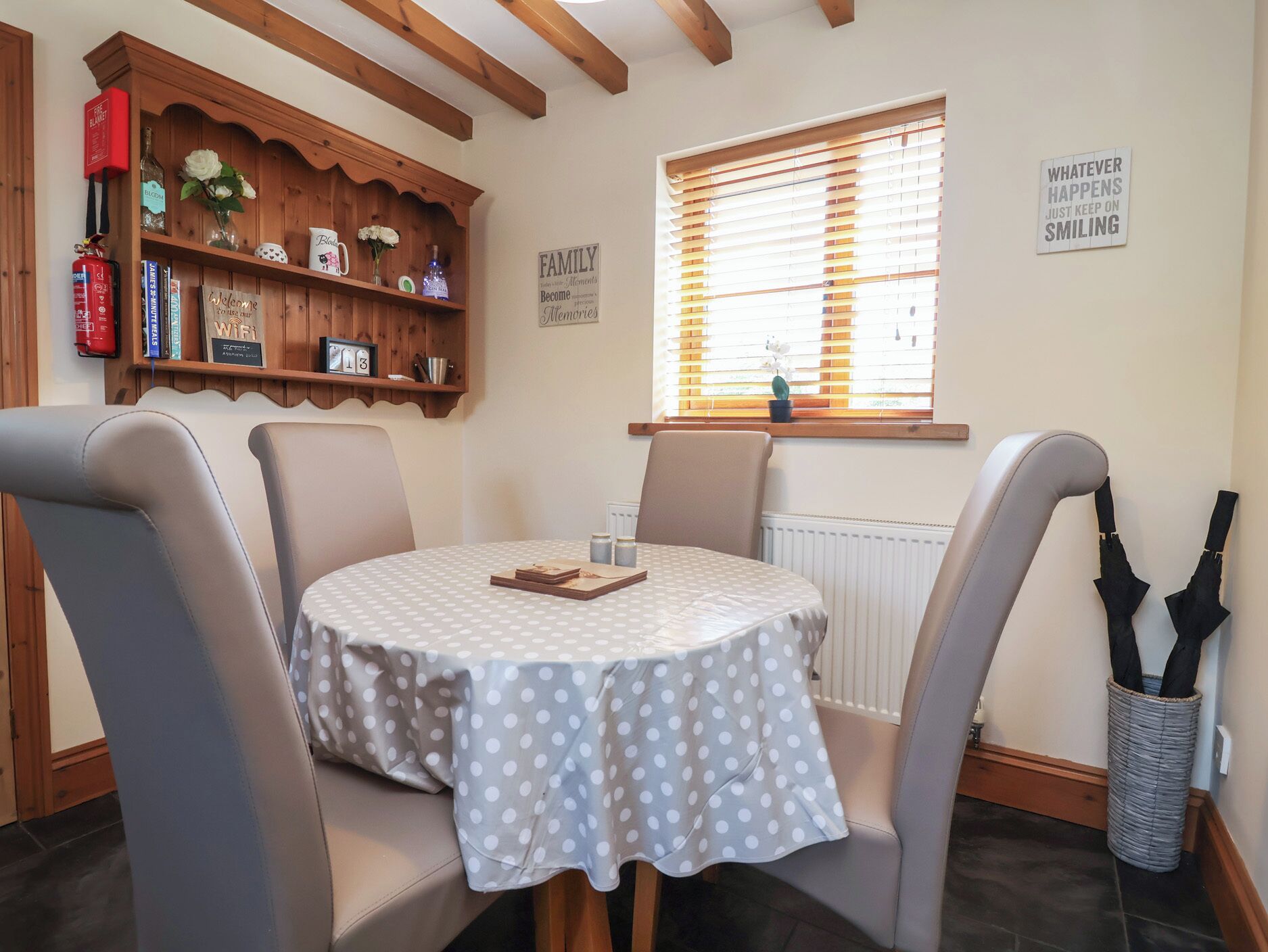 Un comedor con una mesa redonda cubierta por un mantel a lunares, rodeado de sillas, un estante de madera con diversos objetos y una ventana con persianas.