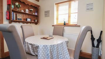 Un comedor con una mesa redonda cubierta por un mantel a lunares, rodeado de sillas, un estante de madera con diversos objetos y una ventana con persianas.