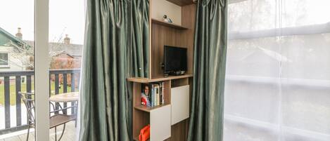 A room with a sliding glass door leading to a balcony, a television on a wooden shelf, and a small table with chairs.