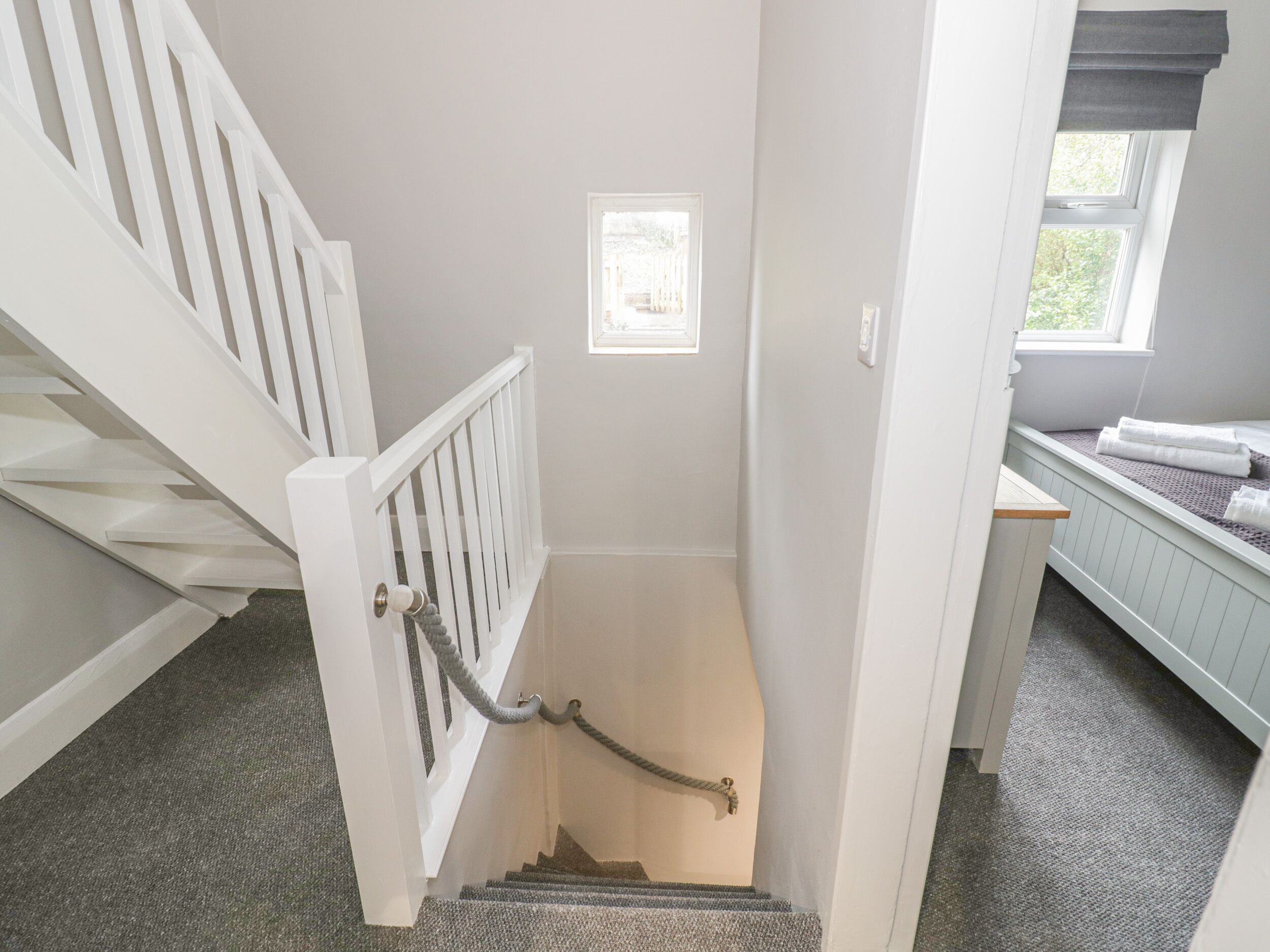 A staircase with a white railing, a small window at the top, and a bed with a gray headboard in the adjacent room.