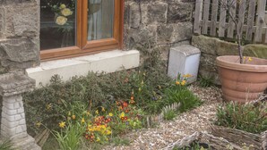 Un bâtiment en pierre doté d’une fenêtre, d’une clôture en bois et d’un jardin où poussent diverses plantes et fleurs.