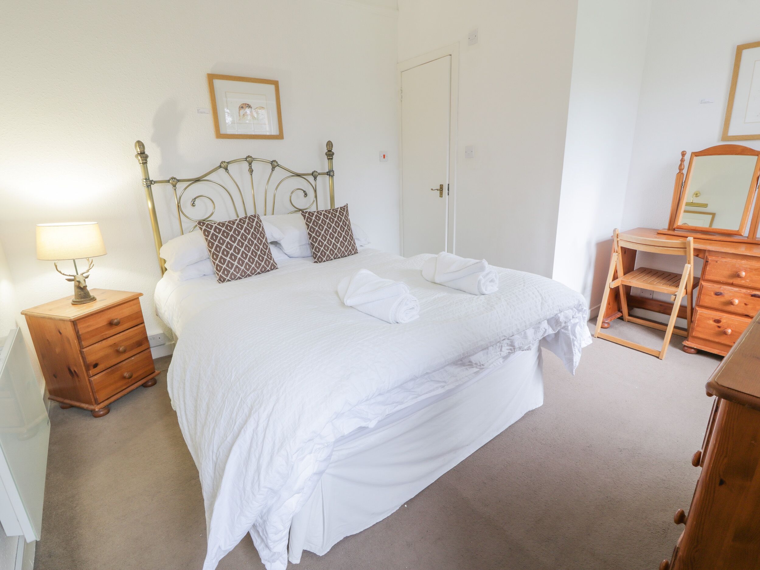 A neatly made bed with white linens and two decorative pillows, a wooden nightstand with a lamp, a wooden dresser with a mirror, and a framed picture on the wall.
