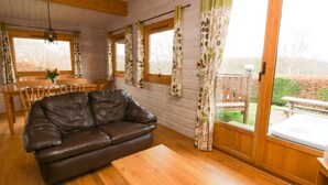 A cozy cabin interior with a leather sofa, wooden dining table, and chairs, large windows with curtains, and a wooden ceiling. - Barn Owl Lodge (Rhayader)