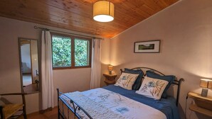 A bedroom with a wooden ceiling, a bed with a floral bedspread, a window with curtains, and a bedside table with a lamp. - Cozy Log Chalet with Terrace, Near Alsace and Ski Resorts (Le Saulcy)