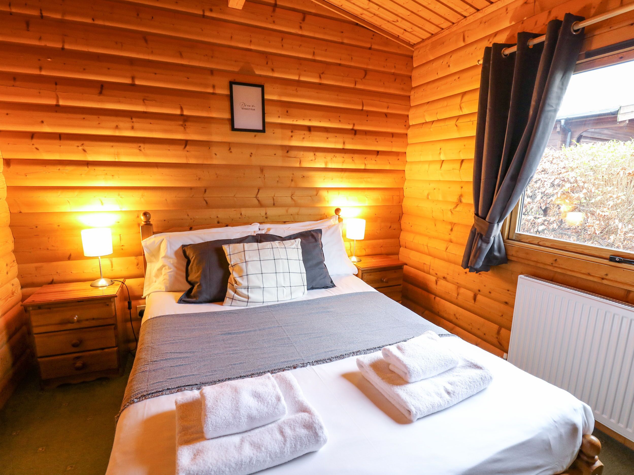 A cozy wooden cabin room with a bed, two bedside lamps, a framed picture on the wall, and a window with curtains.