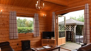 L’intérieur chaleureux d’un chalet, avec un revêtement mural en bois, un téléviseur posé sur un meuble en bois, un canapé en cuir, une petite table et une porte-fenêtre coulissante donnant sur une terrasse extérieure.