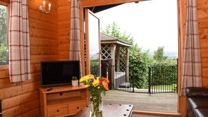 Une cabane en bois avec un téléviseur posé sur un meuble en bois, un vase de fleurs et une vue sur un jardin et des montagnes, visible à travers de grandes fenêtres.