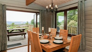 A dining area with a wooden table and chairs, a chandelier, and large windows offering a view of the outdoors. - Mountain View (Rhayader)