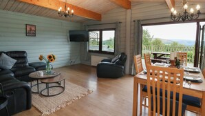 A spacious living room with a wooden ceiling, a black leather sofa, a wooden dining table with chairs, a TV mounted on the wall, and a view of the outdoors through a window. - Mountain View (Rhayader)
