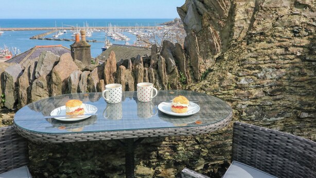 A glass table with two plates of food and two cups, overlooking a harbor with boats and rocky cliffs.
