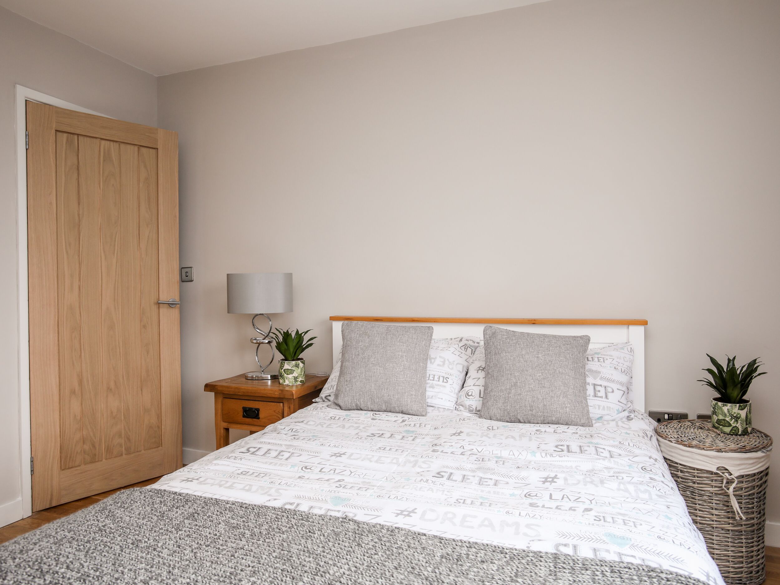Een slaapkamer met een houten deur, een nachtkastje met een lamp en een plant, een bed met beddengoed met dessin, en een rieten mand.