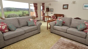 A living room with two grey sofas, patterned cushions, a wooden coffee table, and a view of a green landscape through large windows. - The Yatt (Colwyn Bay)