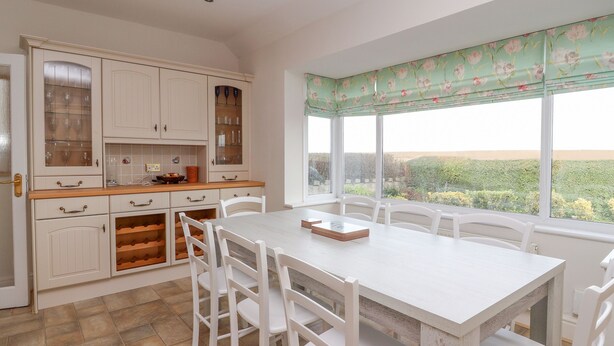 A kitchen with white cabinets, a dining table, and chairs. There is a window with a floral blind. - The Yatt (Colwyn Bay)