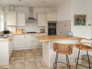 A kitchen with white cabinets, a gas stove, two ovens, and a tiled backsplash. There are two wicker bar stools and a painting on the wall. - The Yatt (Colwyn Bay)