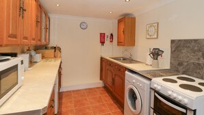 A compact kitchen with wooden cabinets, a microwave, a washing machine, and a stove. - The Old Coach House (Penmaenmawr)