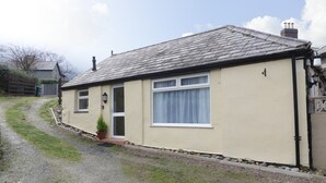 A single-story house with a sloped roof, a front door, and a small garden area with a potted plant. - The Old Coach House (Penmaenmawr)