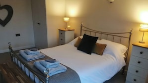 A neatly made bed with pillows, a bedside table with a lamp, a bench, and a wall-mounted heart decoration. - Llofft Yr Yd (Caernarfon)
