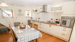 A modern kitchen with wooden cabinets, a built-in oven, and a dining area with a table set for four. - Llofft Yr Yd (Caernarfon)