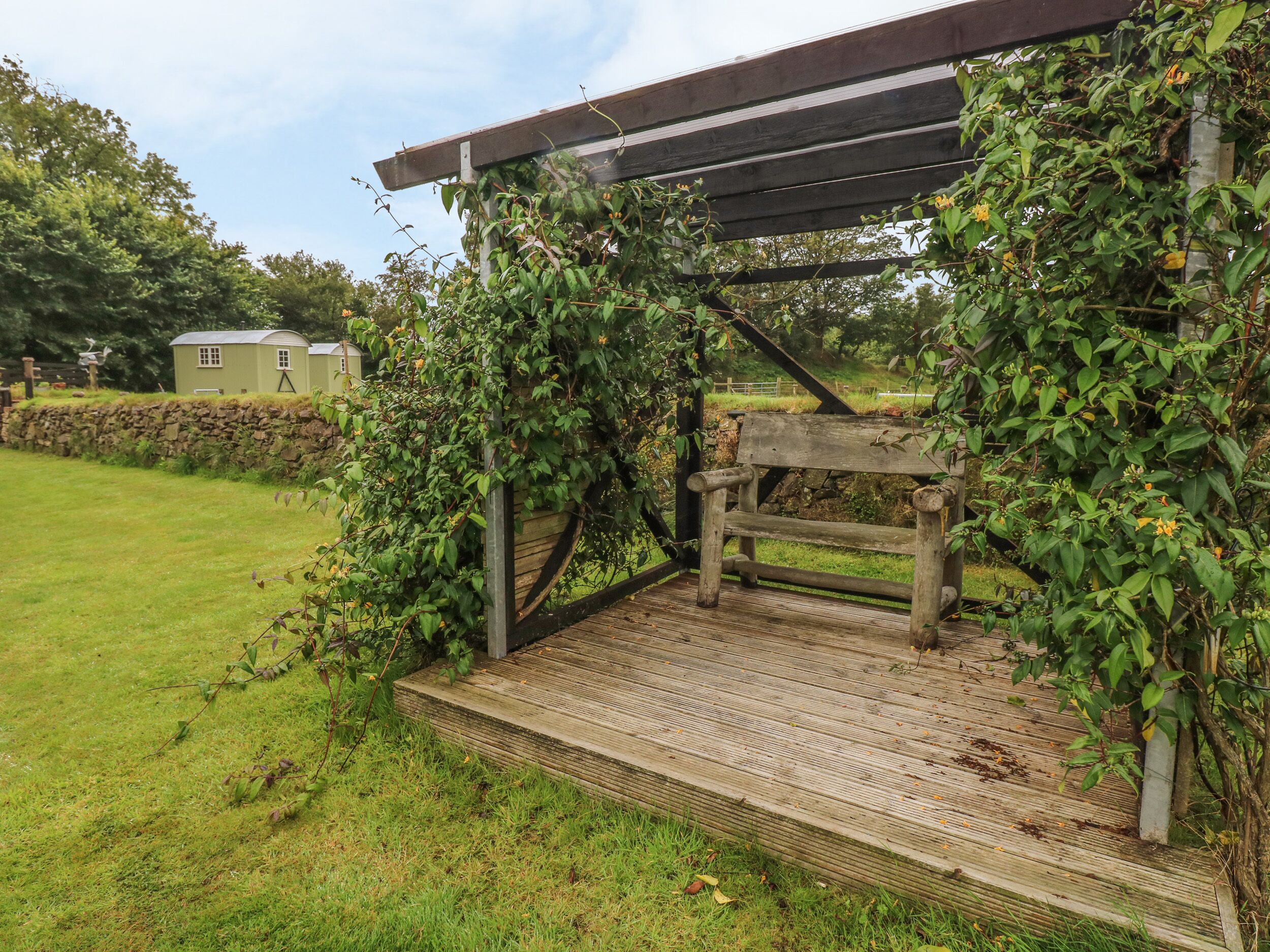 Een houten pergola met een bankje, omringd door weelderige begroeiing en een stenen muur.