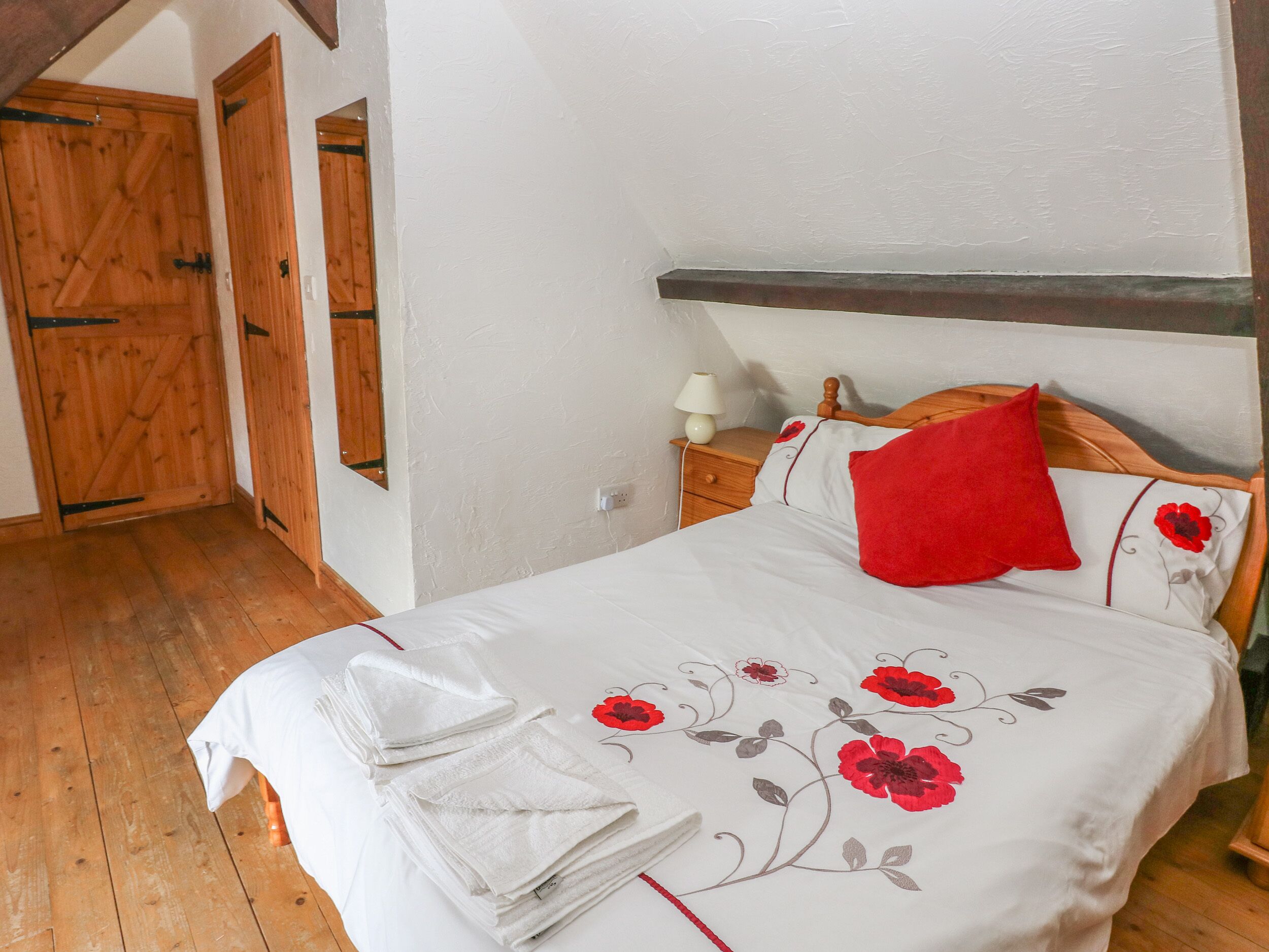 A bedroom with a wooden floor, a bed with a floral bedspread, a red pillow, and a nightstand with a lamp.