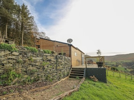 A modern wooden house with a stone retaining wall, a satellite dish, and a deck overlooking a green landscape.
