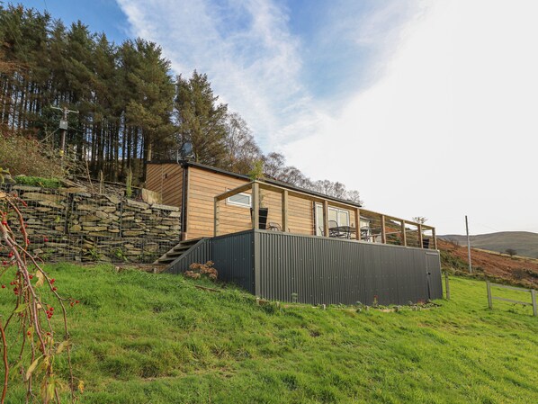 A modern house with a stone retaining wall, a wooden deck, and a view of the surrounding forest. - Haka Lodge (Aberystwyth)
