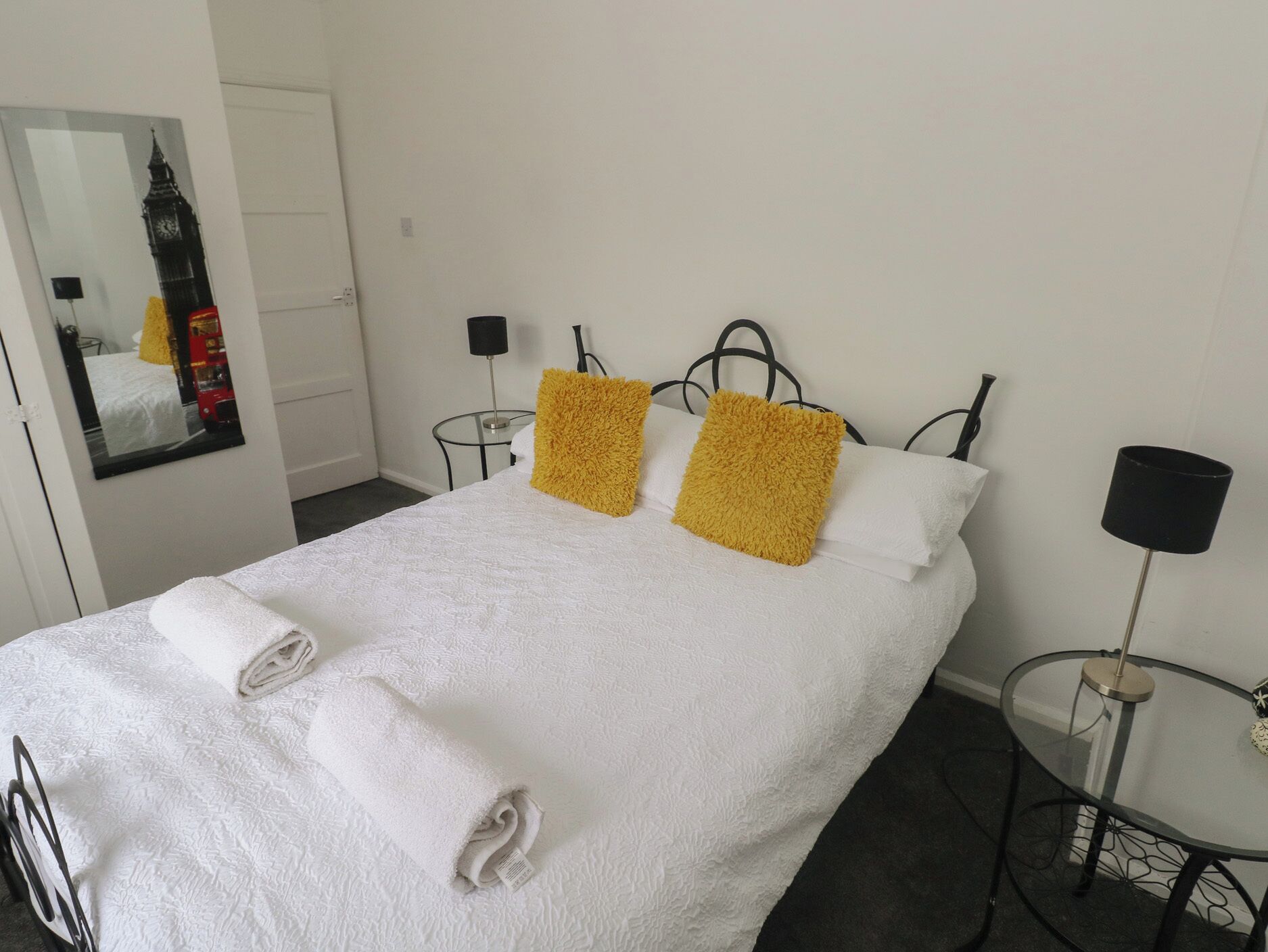 A hotel room with a bed, two pillows, a bedside table, a lamp, and a mirror with a reflection of a red bus.
