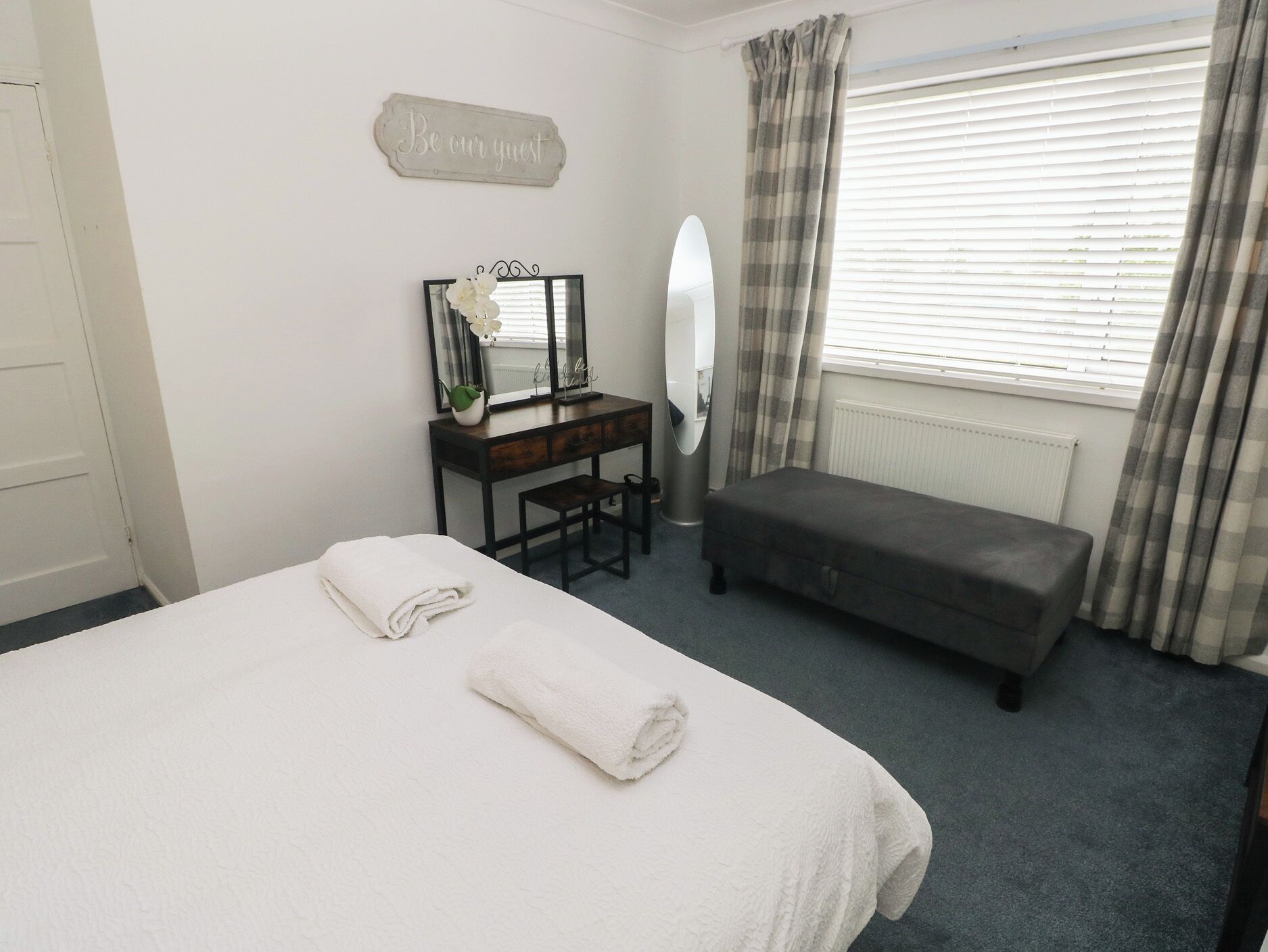 A bedroom with a bed, a desk with a mirror, a bench, and a 'Be our guest' sign.
