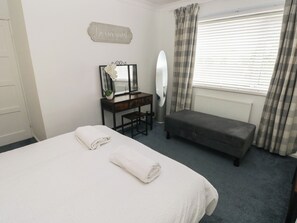 A bedroom with a bed, a desk with a mirror, a bench, and a 'Be our guest' sign. - Cwtch Ar Y Mor (Burry Port)
